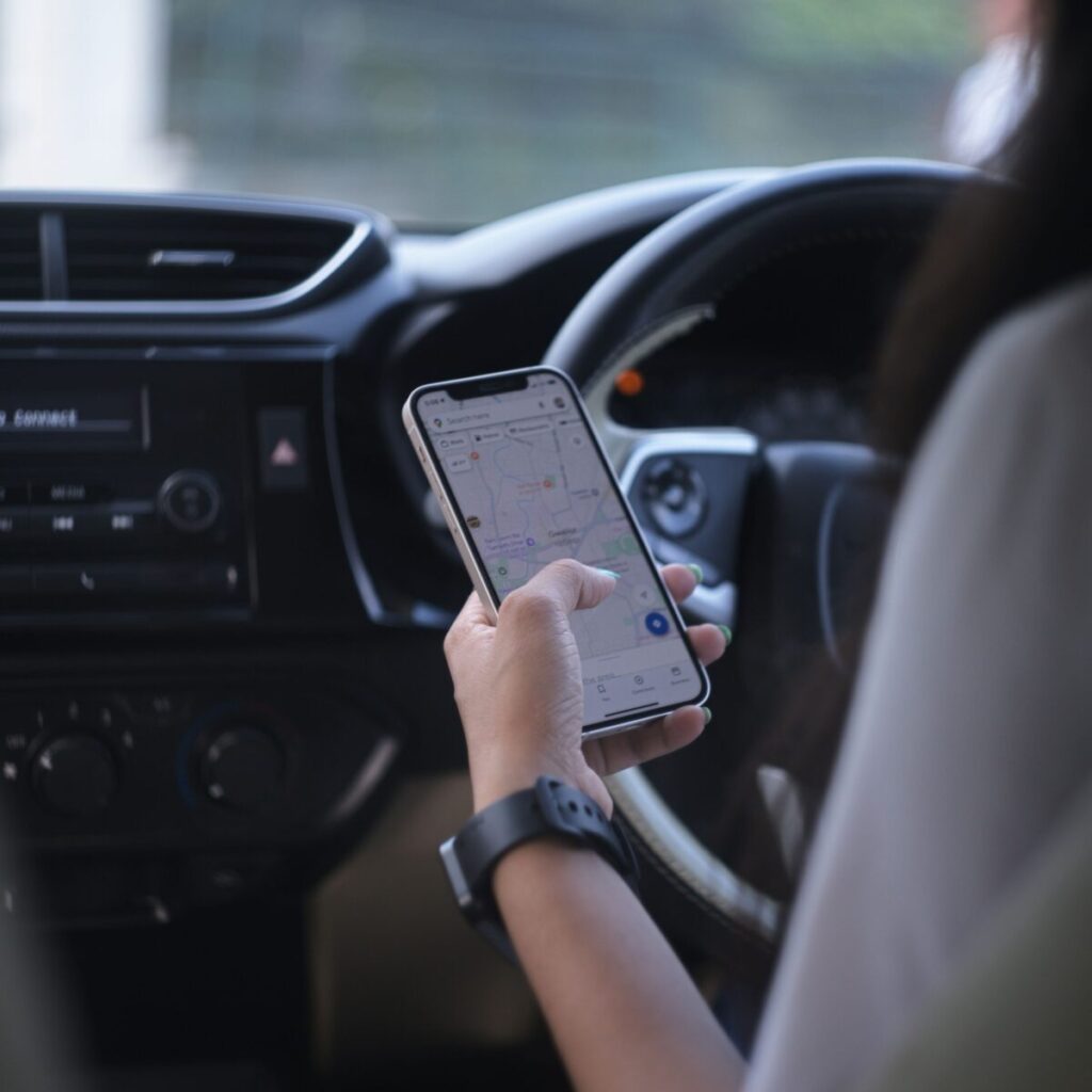 The image shows someone sitting in their car whilst looking at their phone. The phone displays a map of the area.