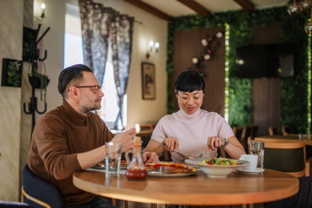 Two tourists sit at a restaurant table in Japan, eating pizza and salad while speaking with each other and practicing basic Japanese phrases for ordering food