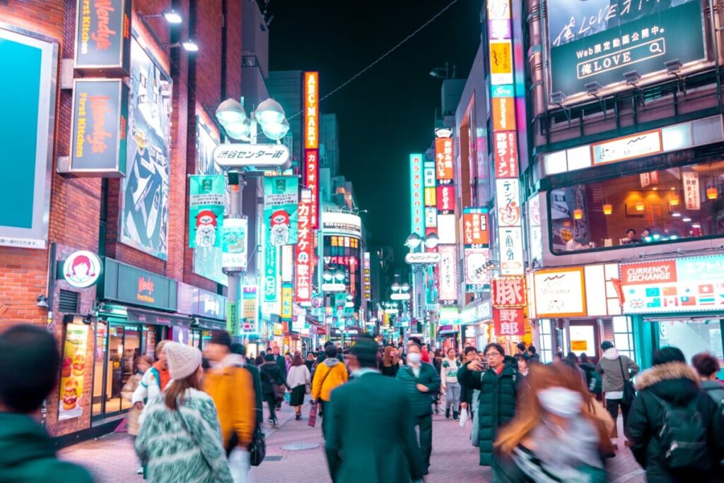 Crowds walking along a neon‑lit shopping street in Shibuya, Tokyo, surrounded by bright shop signs, restaurants and karaoke billboards at night.