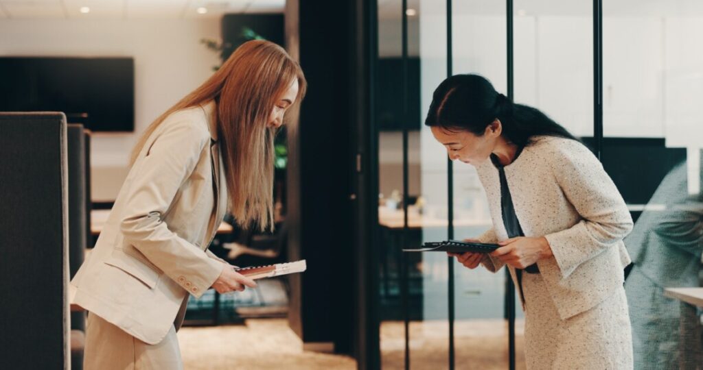 Two women in business attire bow to each other in an office corridor, greeting politely while holding notebooks.